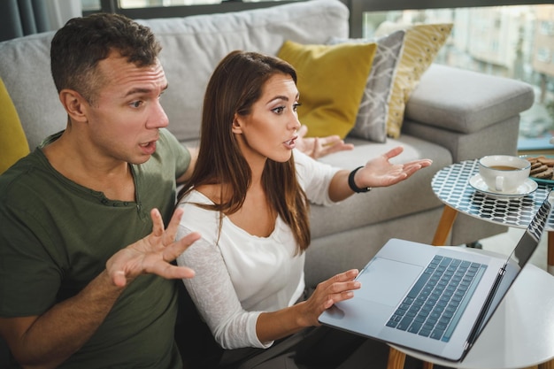 Shot van een mooi stel dat op tapijt in hun woonkamer zit en videogesprekken voert op laptop terwijl ze vrije tijd bij haar thuis heeft tijdens de covid-19-pandemie.