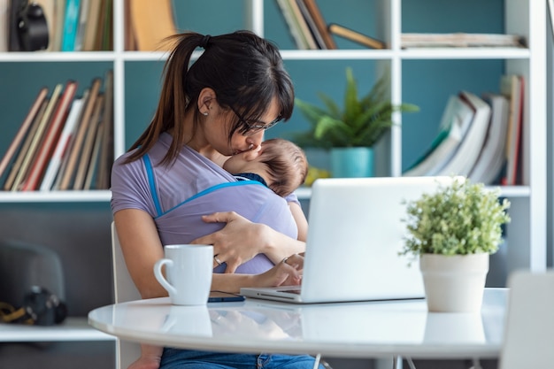 Shot van een mooie jonge moeder die haar baby in een draagdoek kust terwijl ze thuis met een laptop werkt.