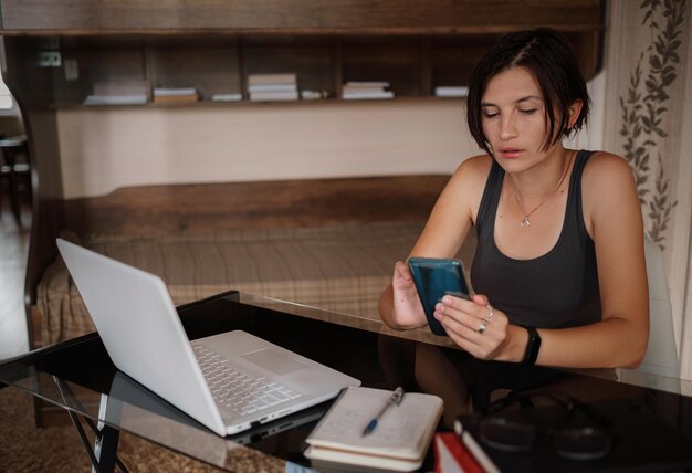 Shot van een mooie jonge vrouw die haar mobiele telefoon gebruikt terwijl ze thuis met een laptop werkt