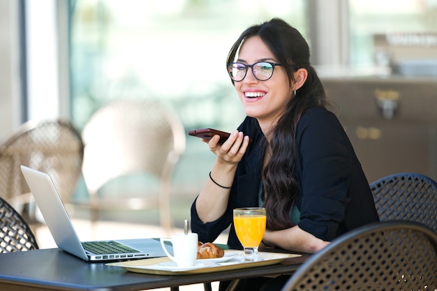 Shot van een mooie jonge vrouw die met haar mobiele telefoon praat en ontbijt terwijl ze met haar laptop in een coffeeshop werkt.