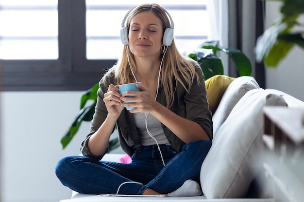 Shot van een mooie jonge vrouw die naar muziek luistert met een koptelefoon terwijl ze thuis een kopje koffie drinkt op de bank.