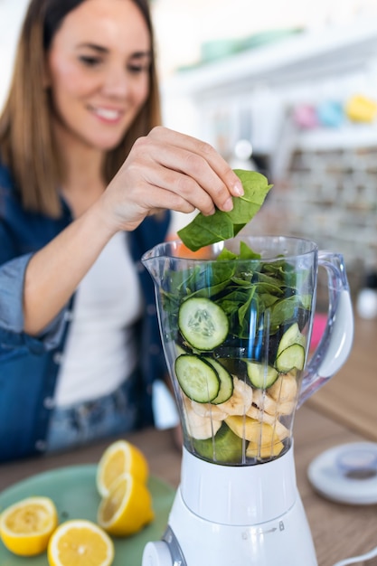 Shot van een mooie jonge vrouw die spinazie in de blender doet voor het bereiden van detox-sap in de keuken thuis.