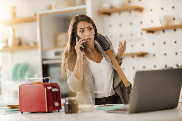 Shot van een multitaskende jonge zakenvrouw die smartphone en laptop in haar keuken gebruikt terwijl ze zich klaarmaakt om naar haar werk te gaan.