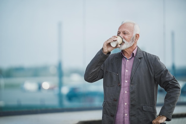 Shot van een senior zakenman die met zijn bagage staat en koffie drinkt buiten de luchthaven.