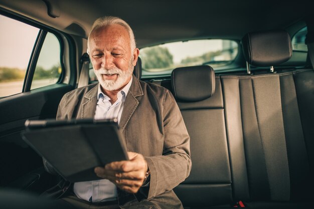 Shot van een succesvolle senior zakenman die een digitale tablet gebruikt terwijl hij op de achterbank van een auto zit tijdens zijn woon-werkverkeer.