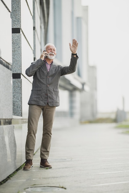 Shot van een succesvolle senior zakenman die op een smartphone praat en iemand begroet terwijl hij even pauzeert voor een bedrijfsgebouw.