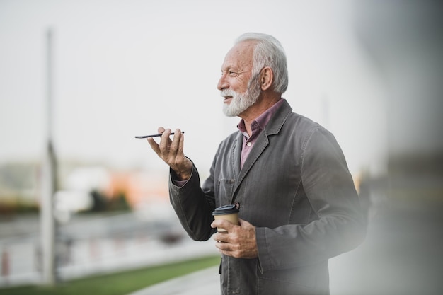 Shot van een succesvolle senior zakenman die via een spraakbericht op zijn smartphone wordt geïnformeerd terwijl hij een koffiepauze houdt voor een bedrijfsgebouw.