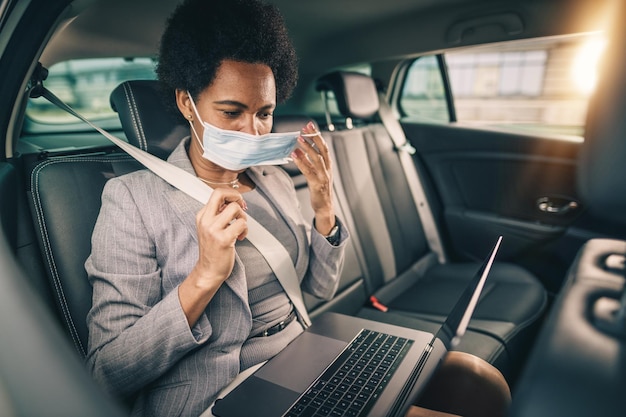 Shot van een succesvolle zwarte zakenvrouw met beschermend masker die een laptop gebruikt terwijl ze op de achterbank van een auto zit tijdens haar woon-werkverkeer tijdens de COVID-19-pandemie.