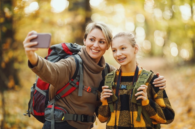 Shot van een tienermeisje en haar moeder die een selfie maken met smartphone tijdens een wandeling samen door het bos in de herfst.