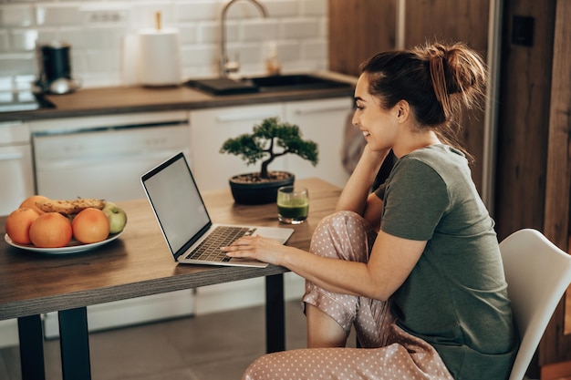 Shot van een vrouw die haar laptop gebruikt en 's ochtends thuis geniet van het ontbijt in haar keuken.