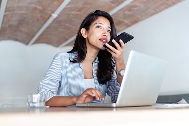 Shot van elegante jonge zakenvrouw praten op mobiele telefoon tijdens het werken met haar laptop op kantoor.