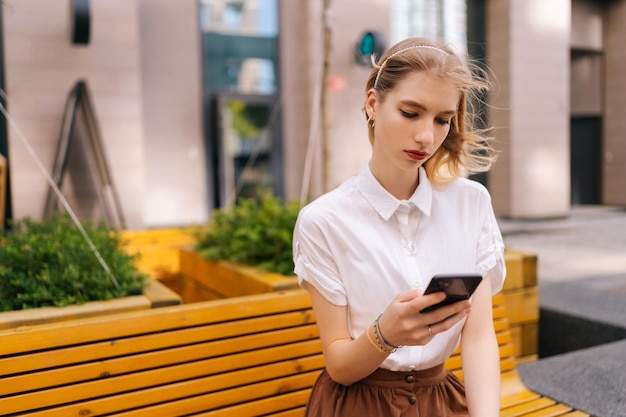 Shot vanuit een gemiddeld perspectief van een serieuze jonge vrouw die een smartphone gebruikt en sms't terwijl ze op een bankje in de straat zit op de achtergrond van een modern gebouw