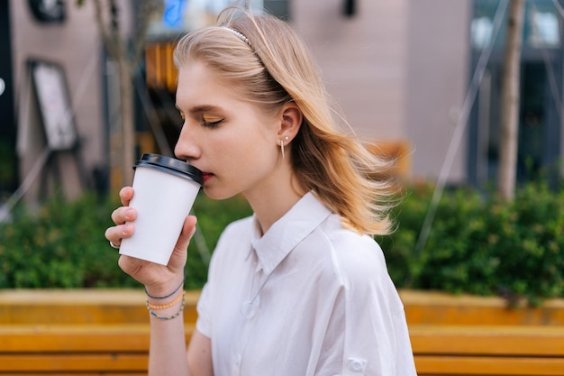 Shot vanuit een gemiddeld perspectief van peinzende jonge vrouw die afhaalkoffie drinkt terwijl ze buiten op iemand wacht in de zomerdag