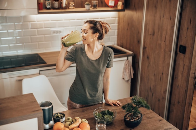 Снимок женщины, пробующей зеленый смузи на кухне дома.