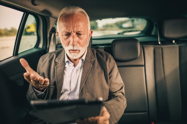 朝の通勤中に車の後部座席に座っているときにデジタルタブレットを使用して心配しているシニアビジネスマンのショット。