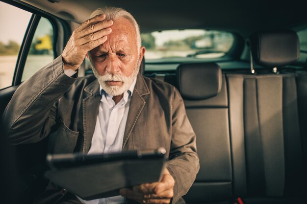 朝の通勤中に車の後部座席に座っているときにデジタルタブレットを使用して心配しているシニアビジネスマンのショット。