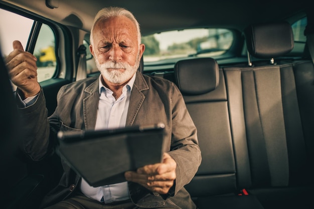 朝の通勤中に車の後部座席に座っているときにデジタルタブレットを使用して心配しているシニアビジネスマンのショット。