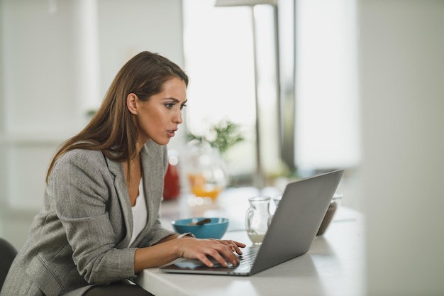 Shot of a young business woman using on her laptop while having a breakfast in the her kitchen during work from home.