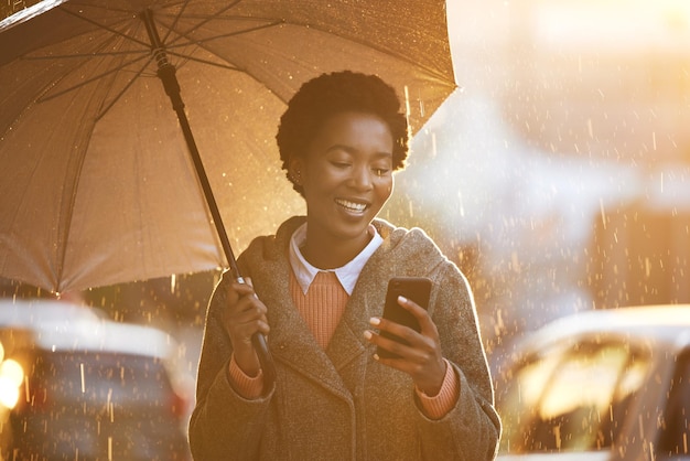 Shot of a young businesswoman using an umbrella and a smartphone while going for a walk in the rain against an urban background