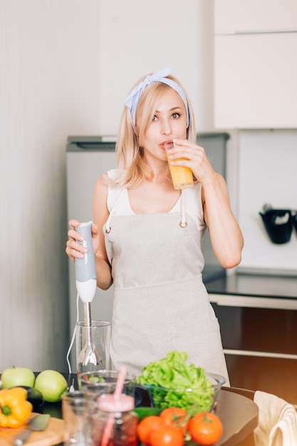Shot of young caucasian woman in apron drinking freshly squeezed juice, holding blender, preparing breakfast, enjoying tasty drink in kitchen at home in morning, looking at camera.