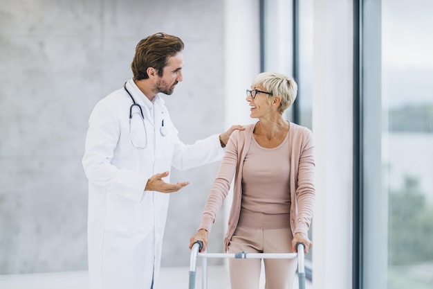 Foto inquadratura di un giovane medico che parla e assiste una paziente anziana a camminare con il suo deambulatore.