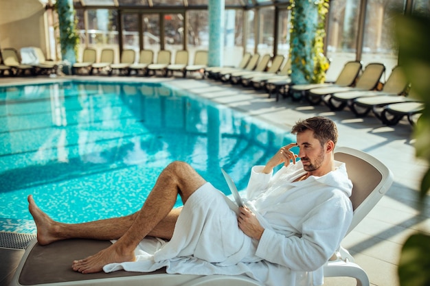 Shot of a young man lying down by the pool and looking at his digital tablet