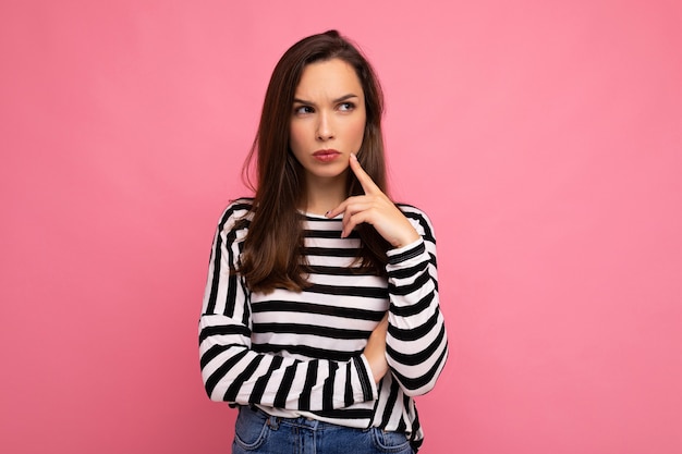 Shot of young sad dissatisfied thoughtful nice cute brunette woman with sincere emotions wearing casual striped sweater isolated on pink background with empty space.