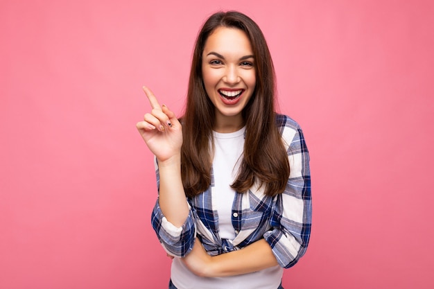 Shot of young smiling happy beautiful brunette woman with sincere emotions wearing trendy check shirt isolated on pink background with empty space and having an idea. Wow concept.