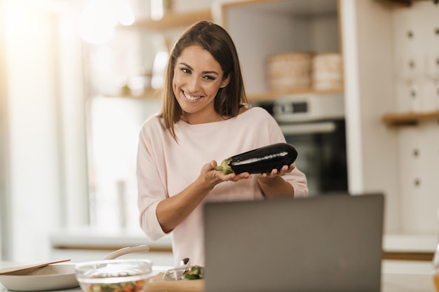 Shot of a young smiling woman preparing a healthy meal and making video blog on a laptop at home.