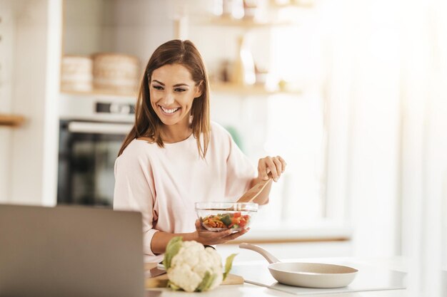 家で健康的な食事を準備しながら、ノートパソコンを使って誰かとビデオ通話をしている笑顔の若い女性のショット。