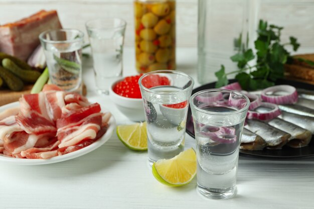 Shots of vodka and tasty snacks on white wooden table, close up