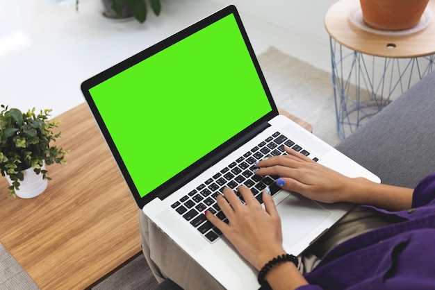 Over shoulder Girl using laptop computer on sofa in living room with blank green screen Work from home concept