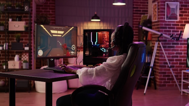 Over shoulder view of african american gamer girl using pc gaming setup playing multiplayer streaming online action game in home living room. Woman in first person shooter tournament talking in headse