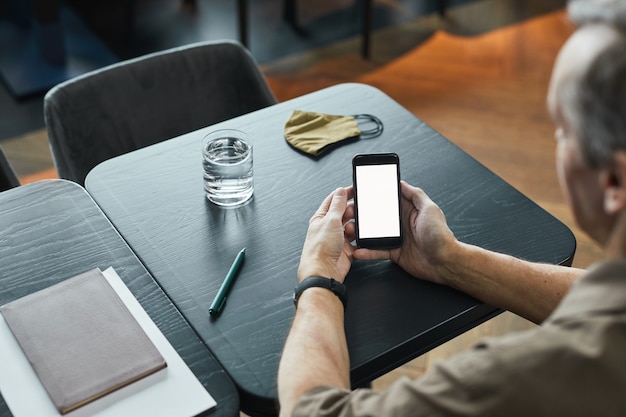 Over shoulder view of aged man sitting at table with glass of water and cloth mask and surfing net on phone, blank screen