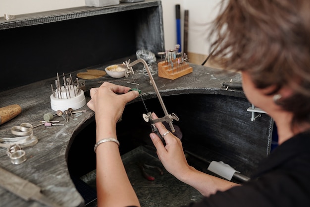 Over shoulder view of female jeweler cutting gemstone with hand jigsaw while making jewelry for sale