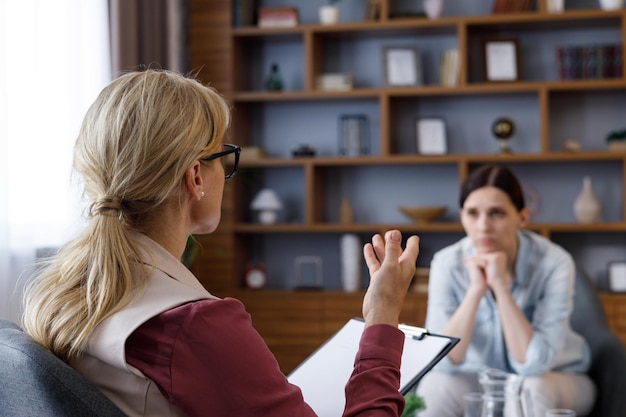 Over shoulder view of female psychologist sitting in armchair talking with upset woman patient Psychologist taking notes on clipboard Psychology mental therapy mental health therapy session