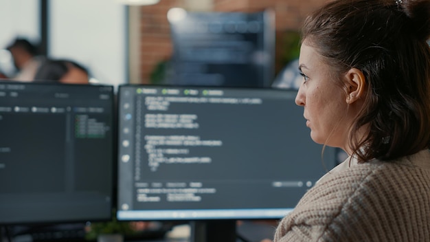 Over shoulder view of focused database designer writing code looking at multiple computer screens displaying algorithm. System engineer coding while colleagues doing teamwork in background.