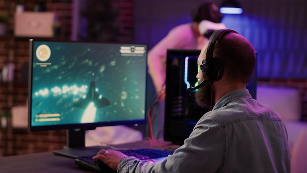 Over shoulder view of gamer playing multiplayer space shooter while gaming girl is fighting in virtual reality in home living room. man streaming action game on pc while gaming girl uses vr goggles.