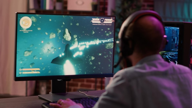 Over shoulder view of gamer streaming fast paced space shooter action game on gaming pc. Man playing multiplayer online tournament talking to team on headset while explaining gameplay to subscribers.