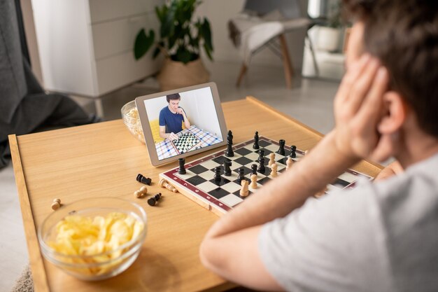 Photo over shoulder view of man sitting at coffee table and deciding about chess move while playing with friend via video chat