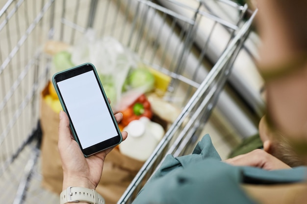 Photo over shoulder view of unrecognizable woman standing at shopping cart and using mobile app while buying food