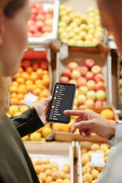 Over shoulder view of young couple checking list of products on smartphone while purchasing food at farmers market