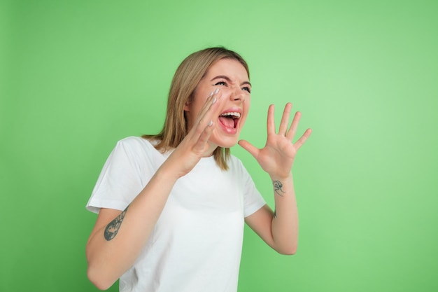 Shouting, screaming. Caucasian young woman's portrait isolated on green  wall. Beautiful female model in white shirt. Concept of human emotions, facial expression, youth.