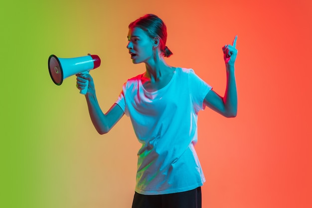 Shouting with megaphone, loudspeaker. Young caucasian girl's portrait on gradient green-orange studio background in neon light. Concept of youth, human emotions, facial expression, sales, ad.