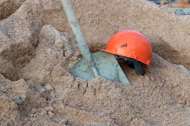 콘크리트를 준비하기 위해 모래 더미에 콘크리트로 덮인 삽과 주황색 건설 노동자의 헬멧
