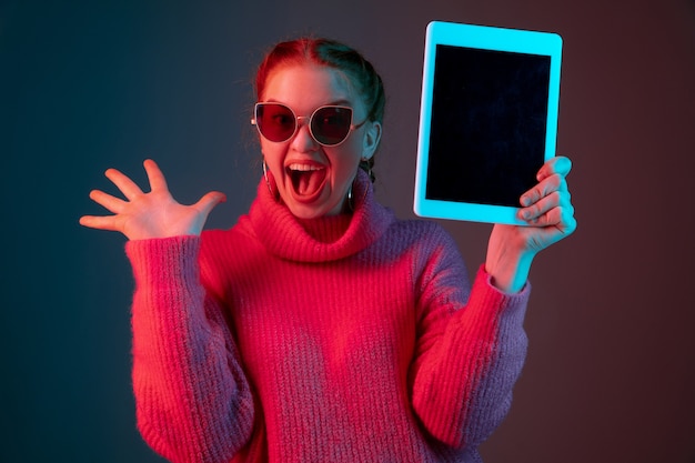 Showing blank tablet screen. Caucasian woman's portrait isolated on gradient studio background in neon light. Beautiful female model with red hair. Concept of human emotions, facial expression, ad.