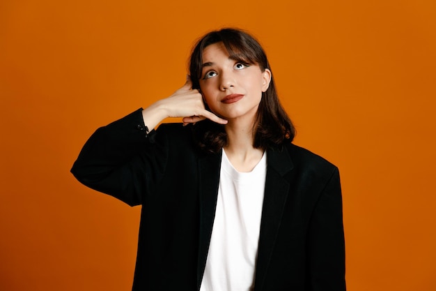 Showing phone call gesture young beautiful female wearing black jacket isolated on orange background