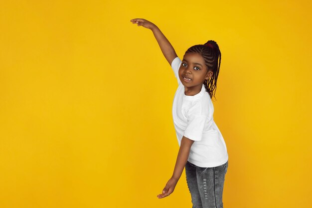 Показывает что-то большое. Портрет маленькой афро-американской девушки на желтом фоне студии. Веселый, красивый малыш. Понятие человеческих эмоций, выражения, продаж, рекламы. Copyspace. Выглядит мило.