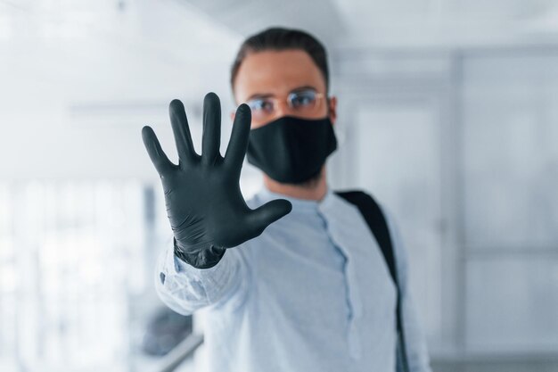 Showing stop gesture by hand In protective mask and gloves Young handsome man in formal clothes indoors in the office at daytime