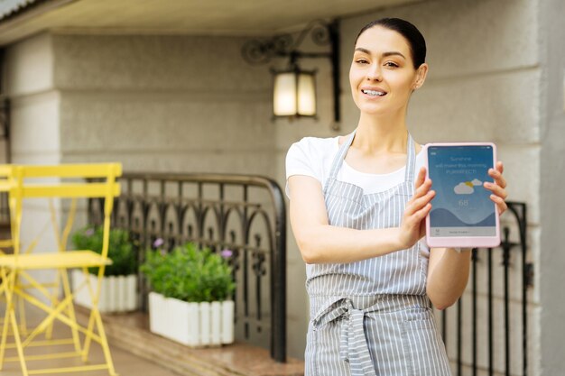 Showing a tablet. Progressive confident waitress smiling and feeling glad while holding a modern device